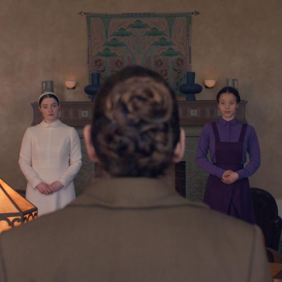 Scene i et rum: to kvinder står med foldede hænder, mens en person i forgrunden ses bagfra.